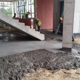 CONTEAM sp. z o.o. - Wylewka betonowa w trakcie realizacji na budowie, widoczni pracownicy w odblaskowych kamizelkach. Surowe wnętrze budynku z betonowymi schodami.