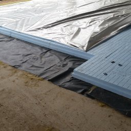 CONTEAM sp. z o.o. - Izolacja podłogowa z płyt styropianowych na czarnej folii, przykryta folią aluminiową. Widoczne spinki mocujące. Etap prac budowlanych.