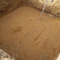 CONTEAM sp. z o.o. - Wykop pod fundament z widocznymi śladami pracy glebogryzarki, częściowo widoczna osoba z łatą pomiarową. Grunt w wykopie jest wyrównany i przygotowany do dalszych prac.