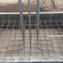 CONTEAM sp. z o.o. - Zbrojenie fundamentu w szalunku z widocznymi pionowymi prętami i siatką zbrojeniową. Detal budowy, betonowa podstawa z elementami stalowymi.