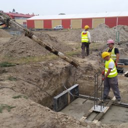 CONTEAM sp. z o.o. - Wylewanie fundamentów pod budynek z użyciem betonomieszarki. Pracownicy w żółtych kamizelkach nadzorują proces. Widoczne zbrojenia i szalunki.