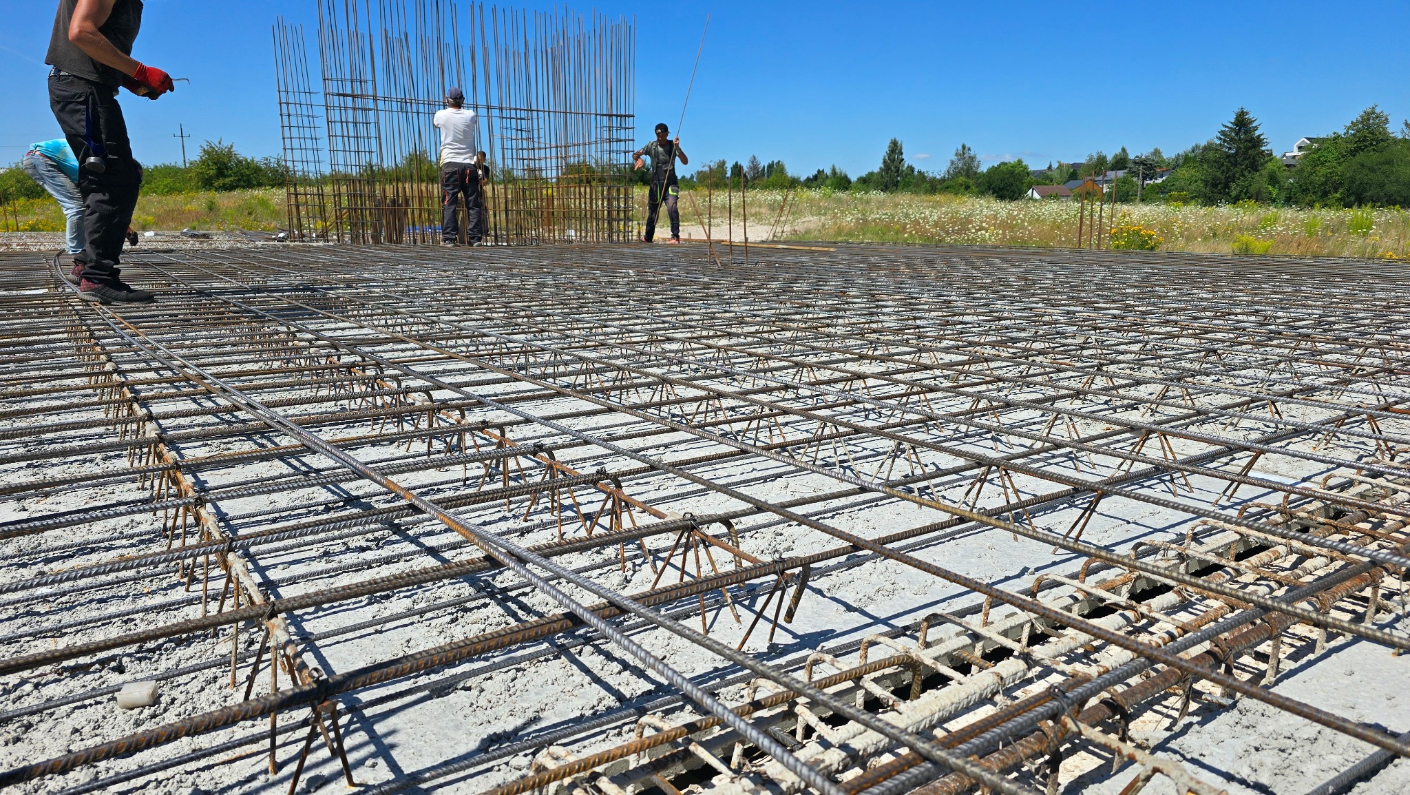 Zbrojenie fundamentów z widocznymi pracownikami na budowie, na tle zieleni i błękitnego nieba. Widok z perspektywy bliskiej ziemi, podkreślający strukturę siatki.