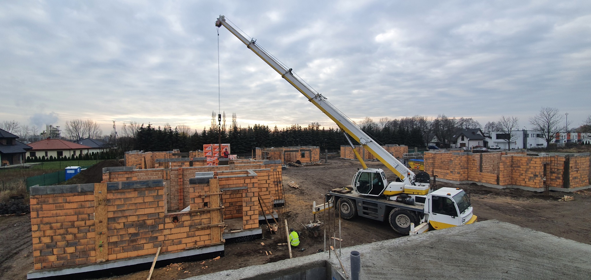 Budowa osiedla domów z cegły ceramicznej. Na pierwszym planie dźwig samochodowy podnosi paletę z bloczkami. W tle widoczne zarysy kolejnych budynków.