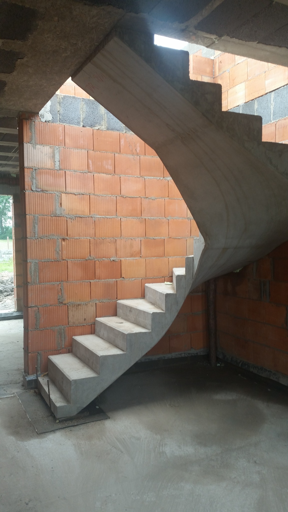 Surowe, betonowe schody spiralne w trakcie budowy domu z czerwonej cegły. Widoczne niedokończone ściany i surowy beton. Minimalistyczny design w stanie surowym.