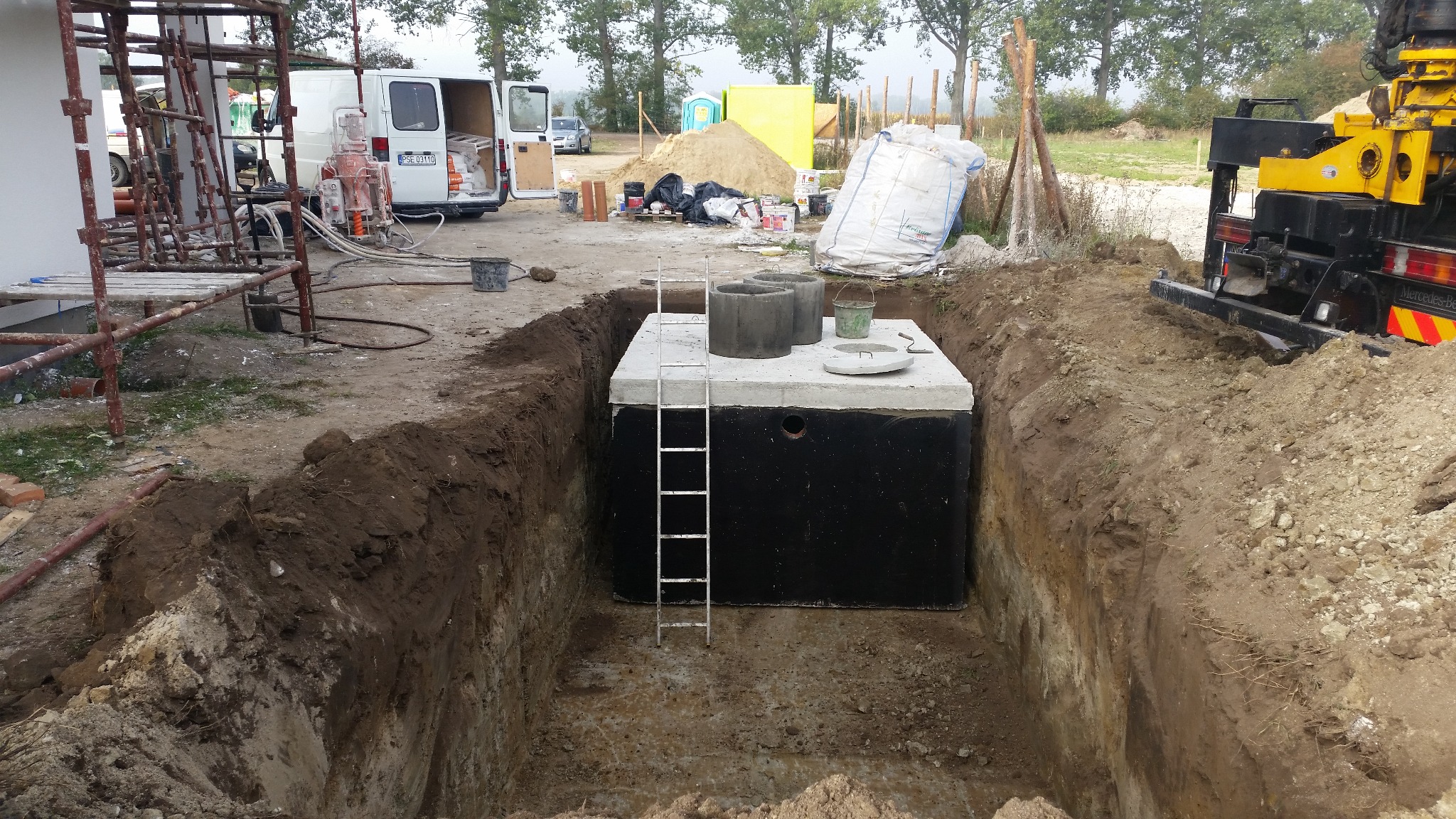 Montaż betonowego zbiornika w wykopie, widoczna drabinka, elementy betonowe na górze. W tle pojazd budowlany i materiały budowlane.