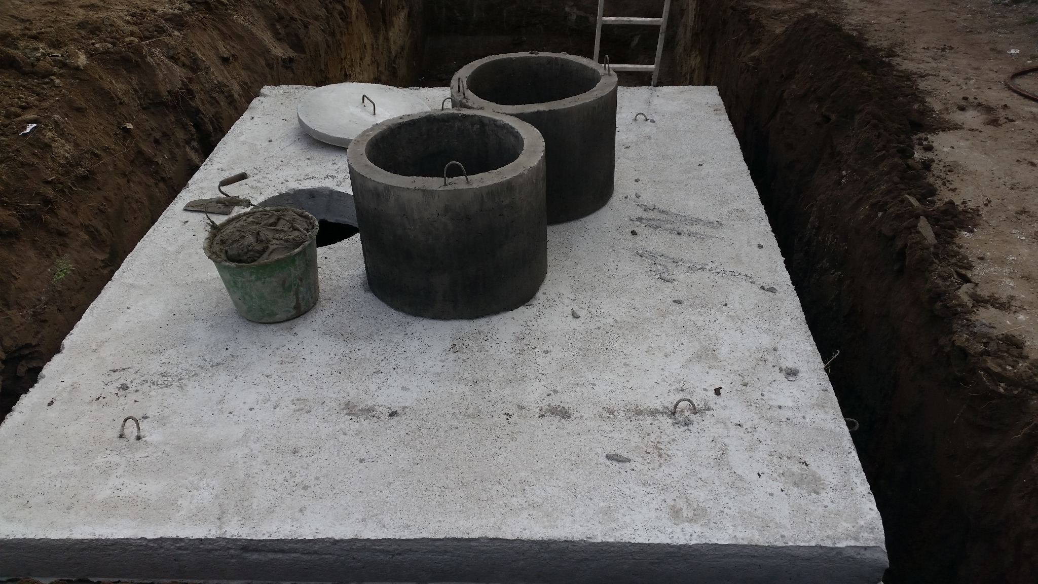 Betonowa płyta z dwoma studzienkami i wiadrem zaprawy w wykopie. Widoczna drabina. Elementy betonowe z metalowymi uchwytami.