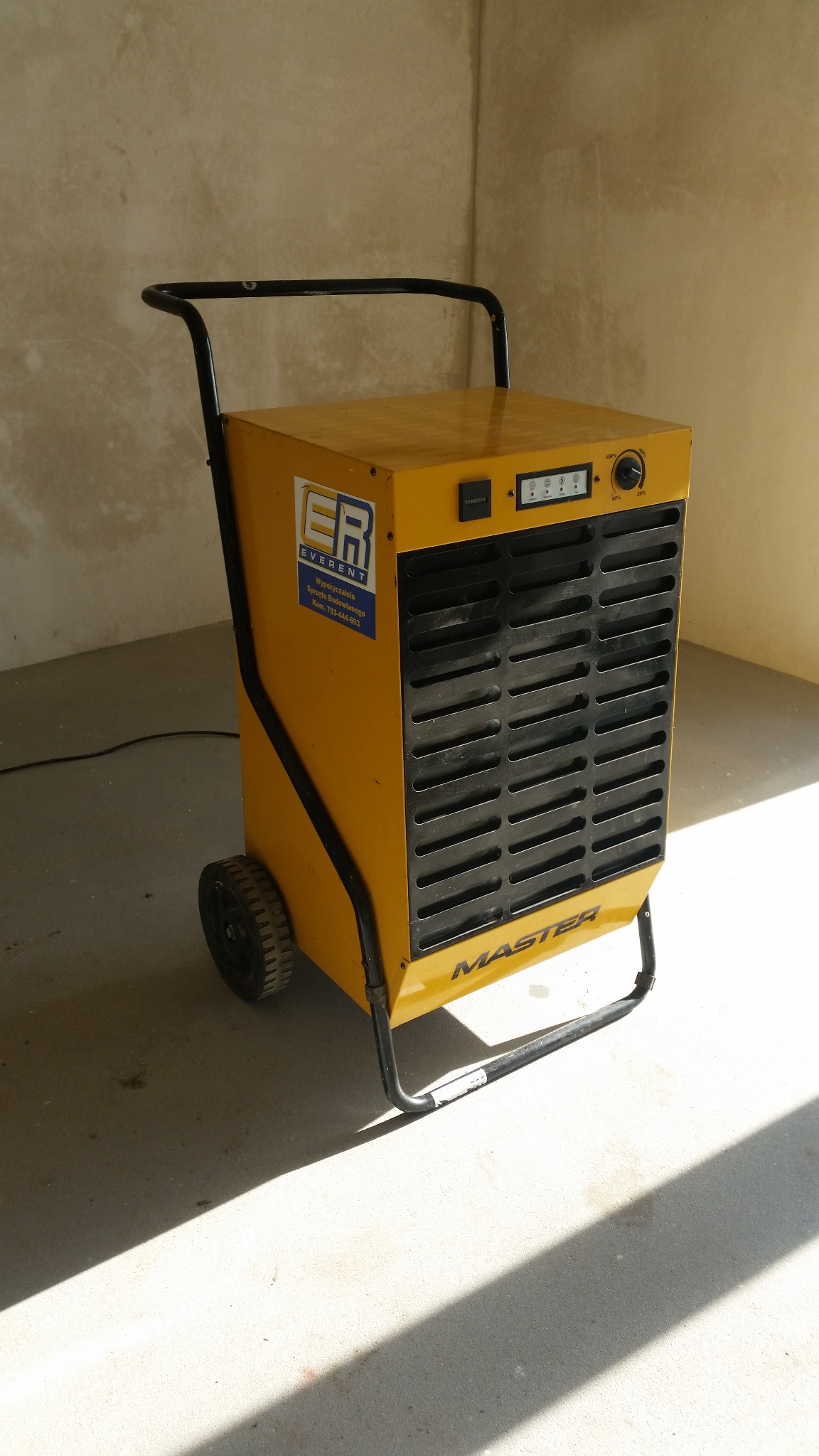 Żółty osuszacz powietrza Master na kółkach, z czarną kratką i uchwytem, stoi w rogu pomieszczenia z surowymi ścianami. Widoczna naklejka firmy Everent.