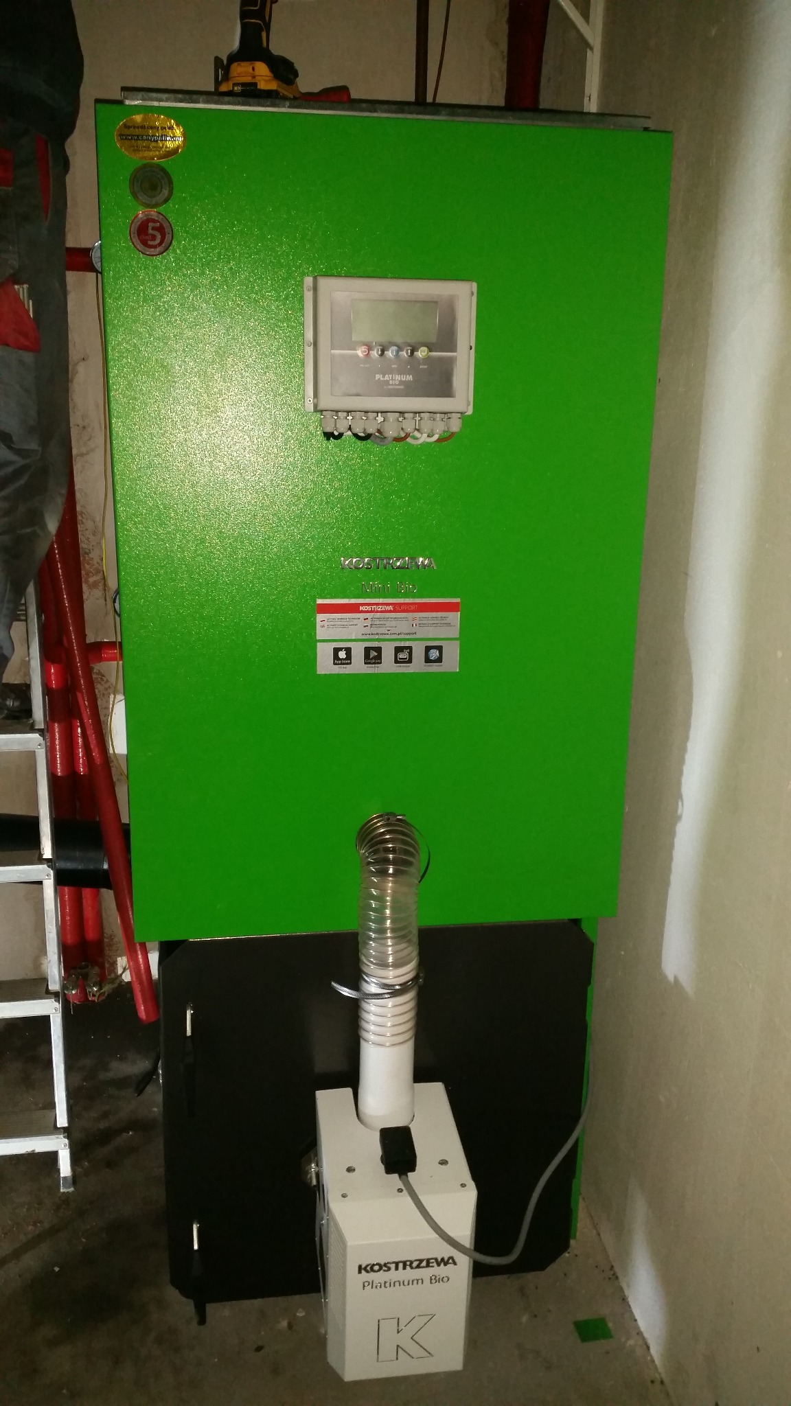 Zielony piec Kostrzewa Platinum Bio z podłączonym przewodem, widoczny panel sterowania i naklejki informacyjne. Obok drabina i fragment instalacji. Ściana w tle.