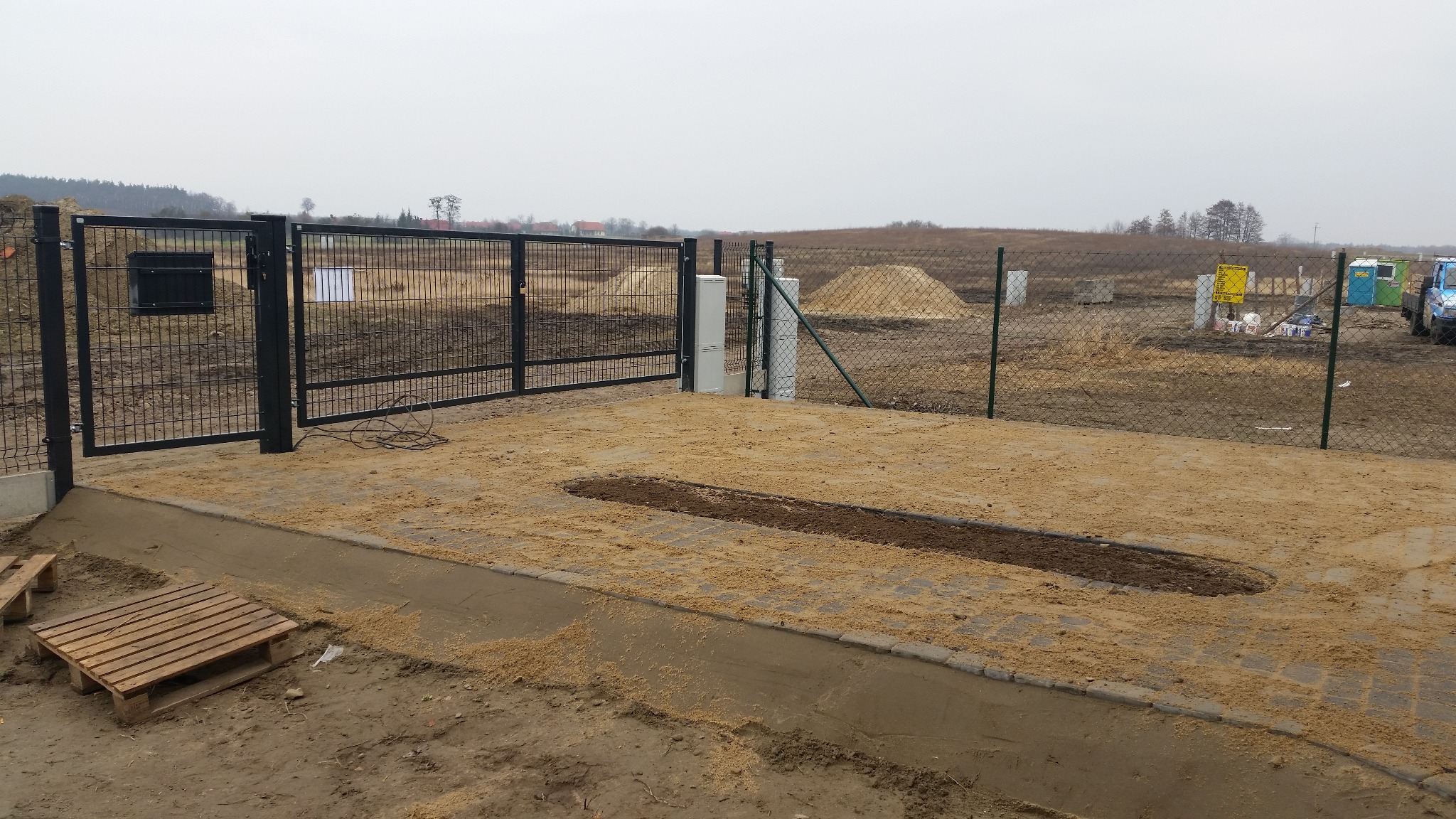 Montaż bramy wjazdowej z czarnej siatki, widoczne świeżo ułożone kostki brukowe posypane piaskiem. W tle teren budowy z hałdami piasku i przenośną toaletą.