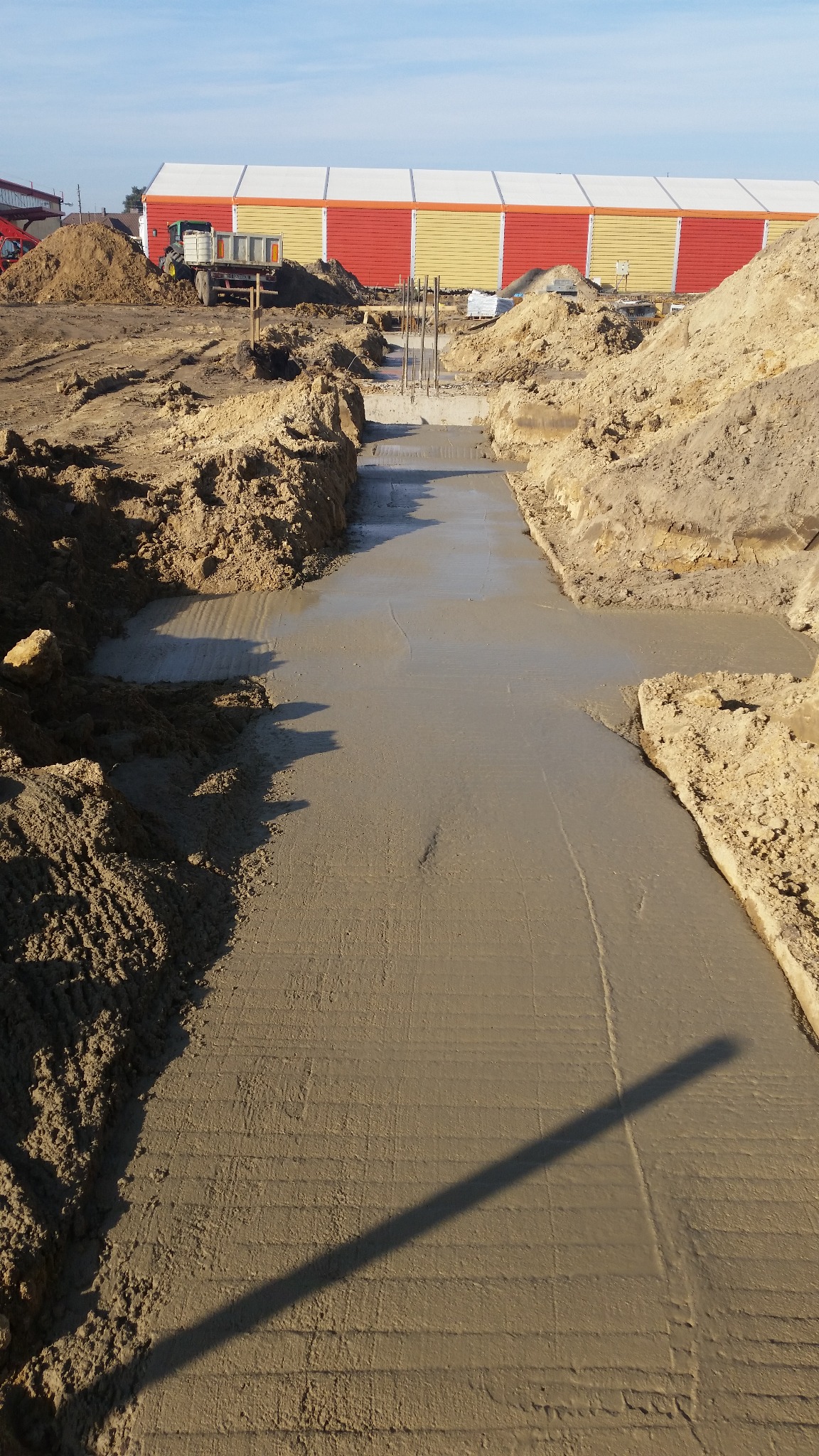 Wylany fundament pod budynek z wystającymi prętami zbrojeniowymi, otoczony wykopem i ziemią. W tle widoczny budynek z żółto-czerwonymi panelami.