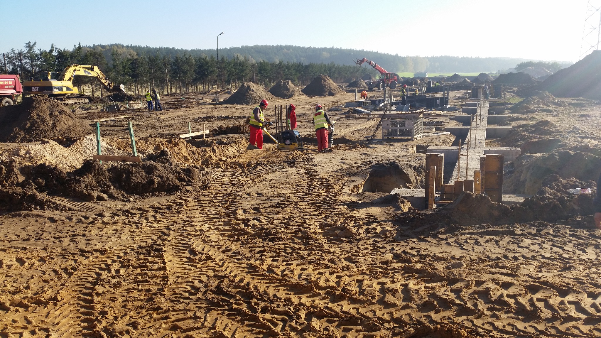 Fundamenty pod budowę w Poznaniu: pracownicy w odblaskowych kamizelkach, wykop, maszyny budowlane, zbrojenia i szalunki w słoneczny dzień.