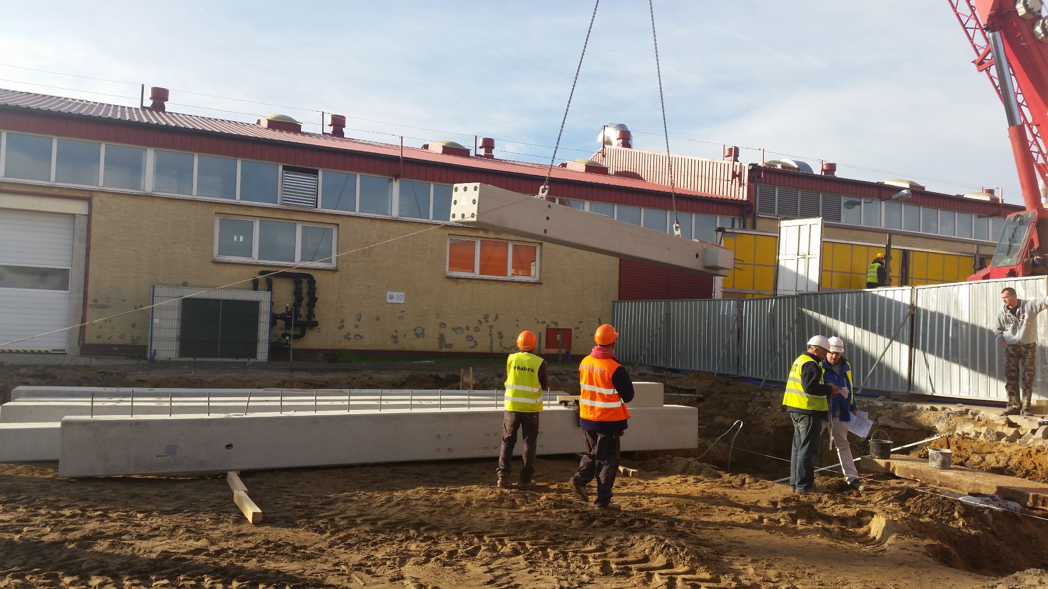 Montaż żelbetowego elementu konstrukcyjnego hali przez dźwig. Na budowie pracownicy w kaskach i kamizelkach odblaskowych. W tle budynek przemysłowy z czerwonym dachem.