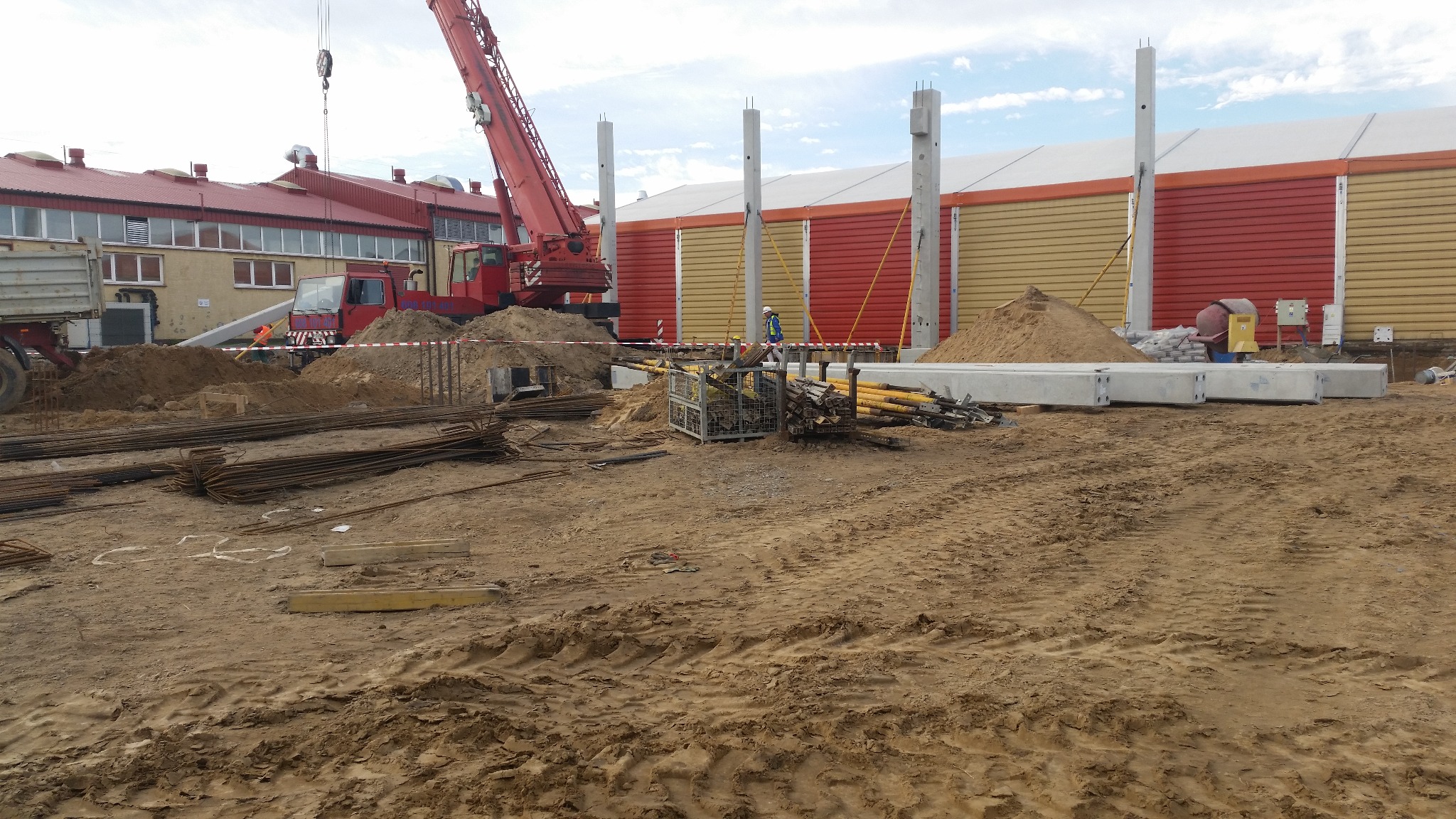 Budowa hali: czerwony dźwig, betonowe słupy i elementy konstrukcyjne na placu budowy. W tle budynek z żółto-czerwonymi panelami.