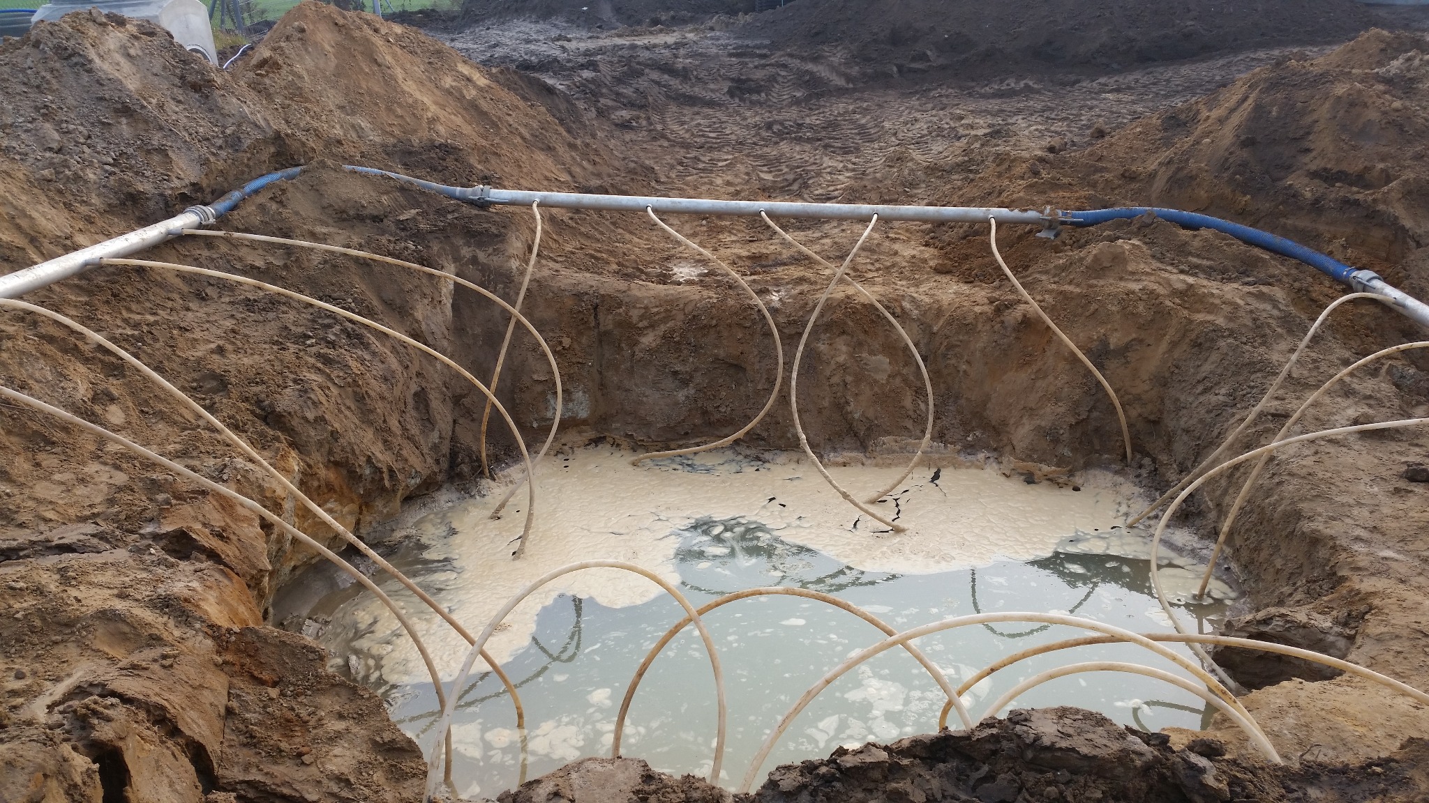 Odwadnianie wykopu pod budowę: system rur zanurzonych w wodzie, podłączonych do głównej rury odprowadzającej wodę. Grunt gliniasty, widoczne ślady po wykopie.