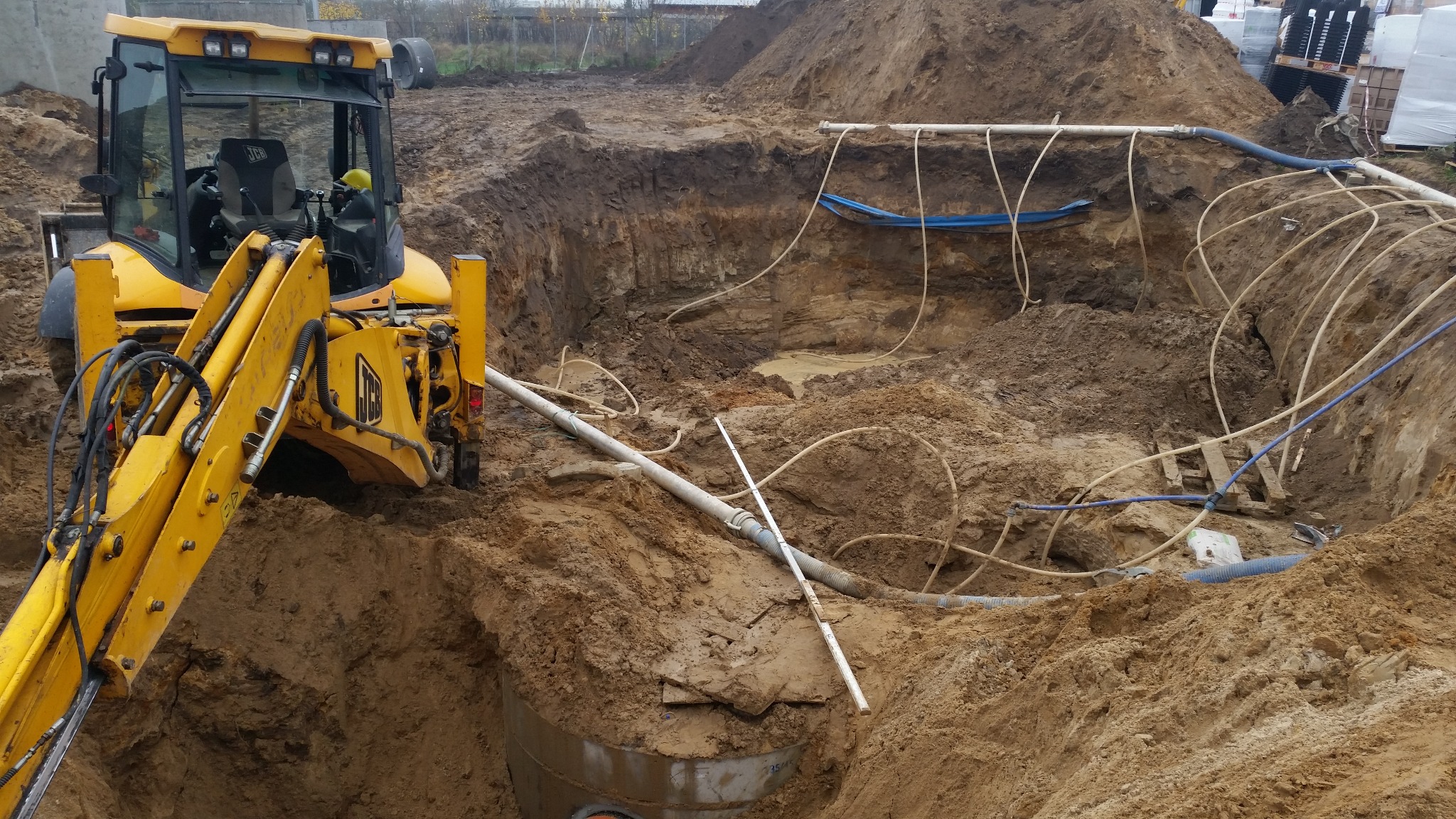Żółta koparka przy wykopie z rurami i kablami. Widoczne warstwy ziemi i elementy konstrukcyjne. Praca w toku, przygotowanie terenu pod budowę.