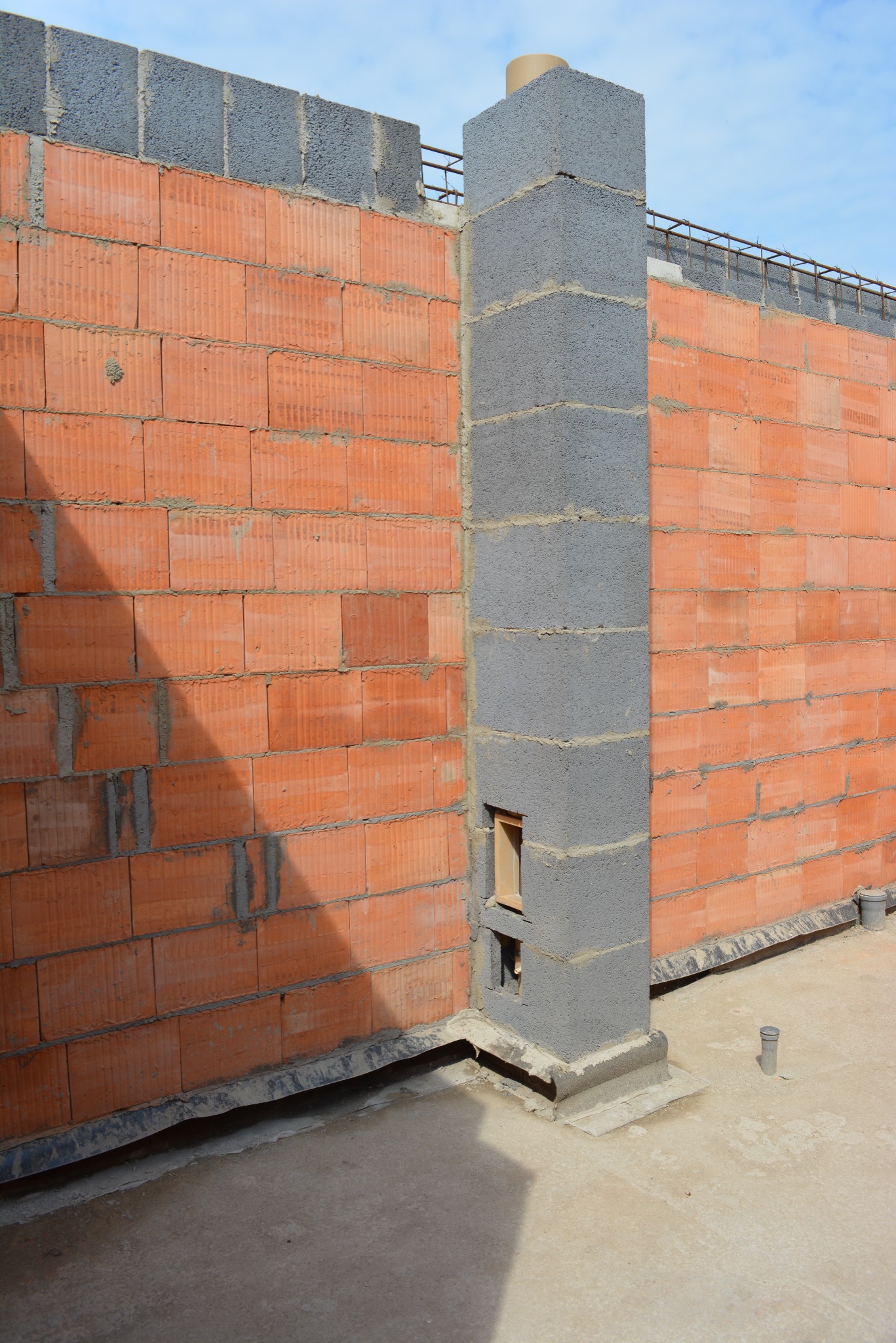 Wznoszenie ściany z czerwonej cegły ceramicznej i szarego słupa wentylacyjnego z pustaków betonowych. Widoczne zbrojenie wieńca i fragment nieba.