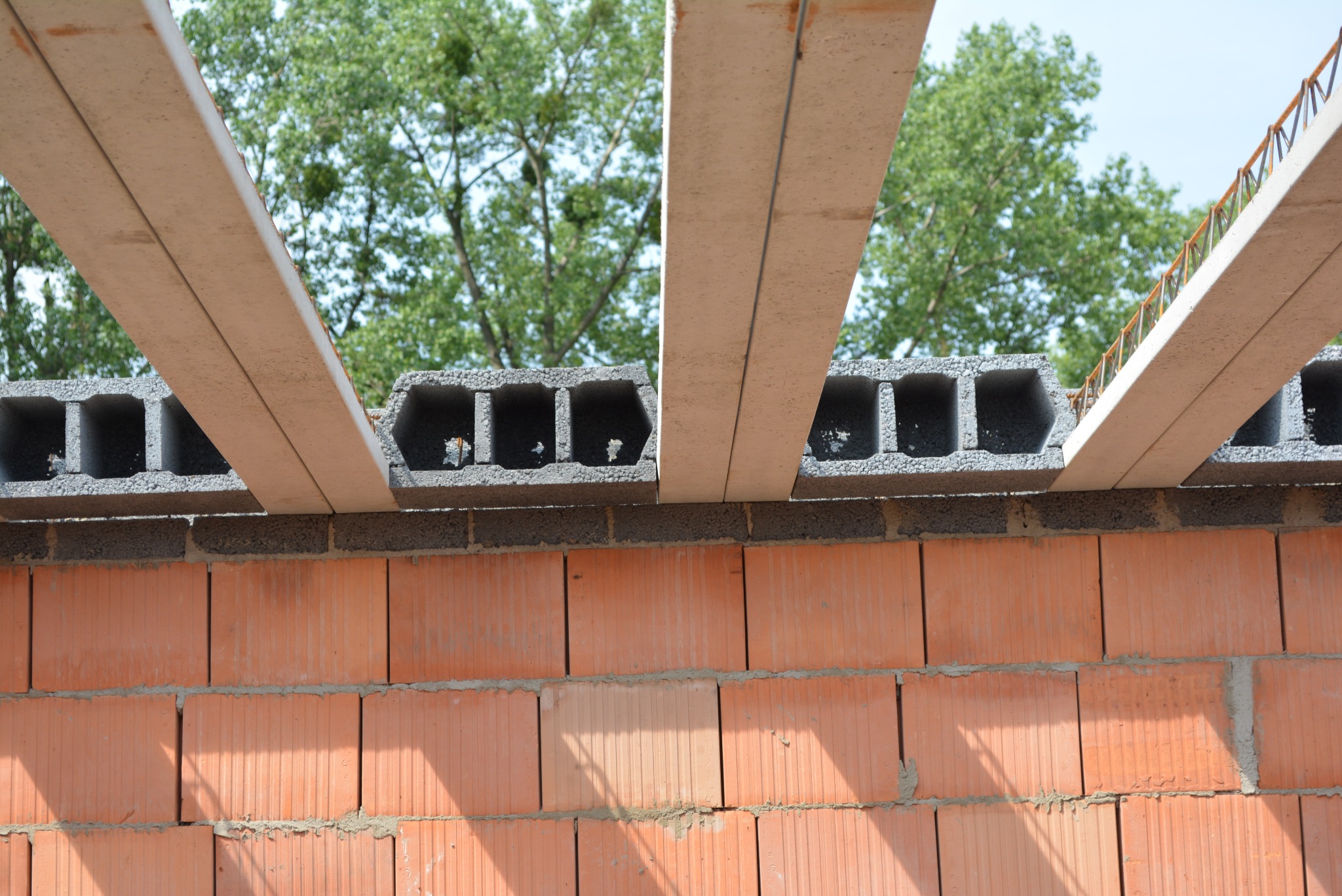 Wznoszenie ściany z czerwonej cegły ceramicznej, z widocznymi szarymi bloczkami betonowymi i elementami stropu Teriva, zieleń drzew w tle.