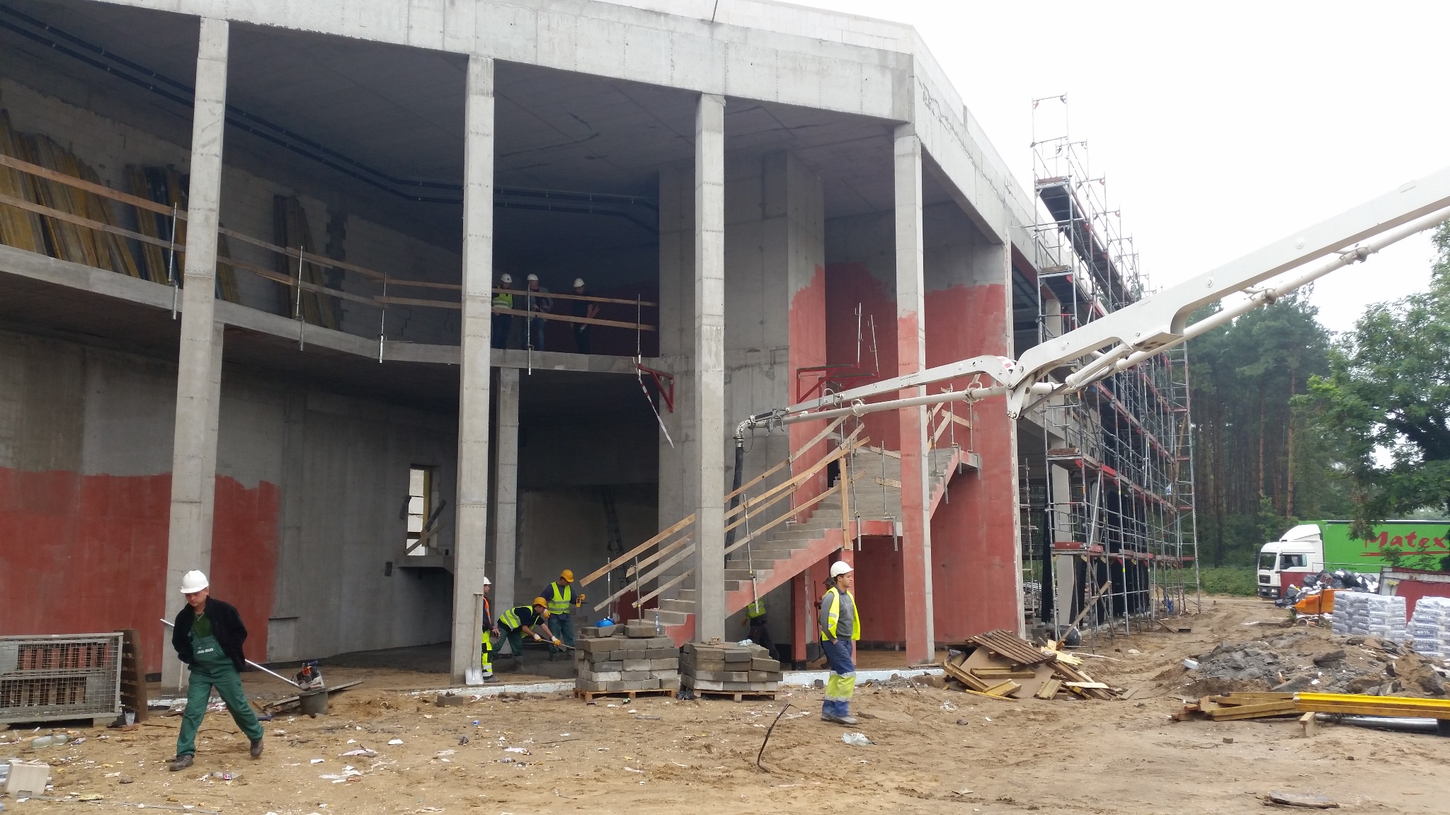 Budowa budynku żelbetowego z widocznym szalunkiem, betonowymi kolumnami i robotnikami w kaskach. Na pierwszym planie pompa do betonu i materiały budowlane.