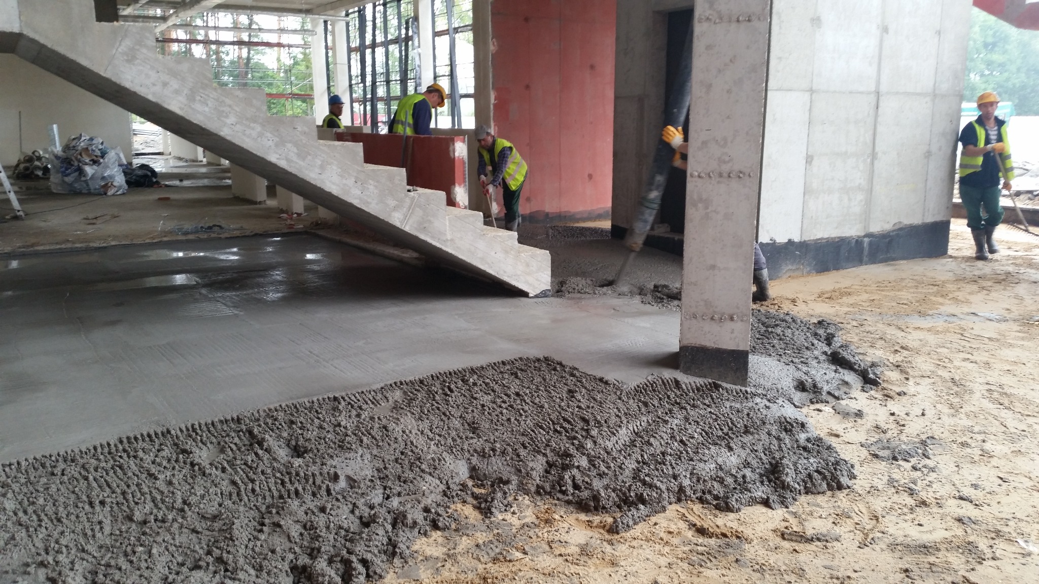 Wylewka betonowa w trakcie realizacji na budowie, widoczni pracownicy w odblaskowych kamizelkach. Surowe wnętrze budynku z betonowymi schodami.