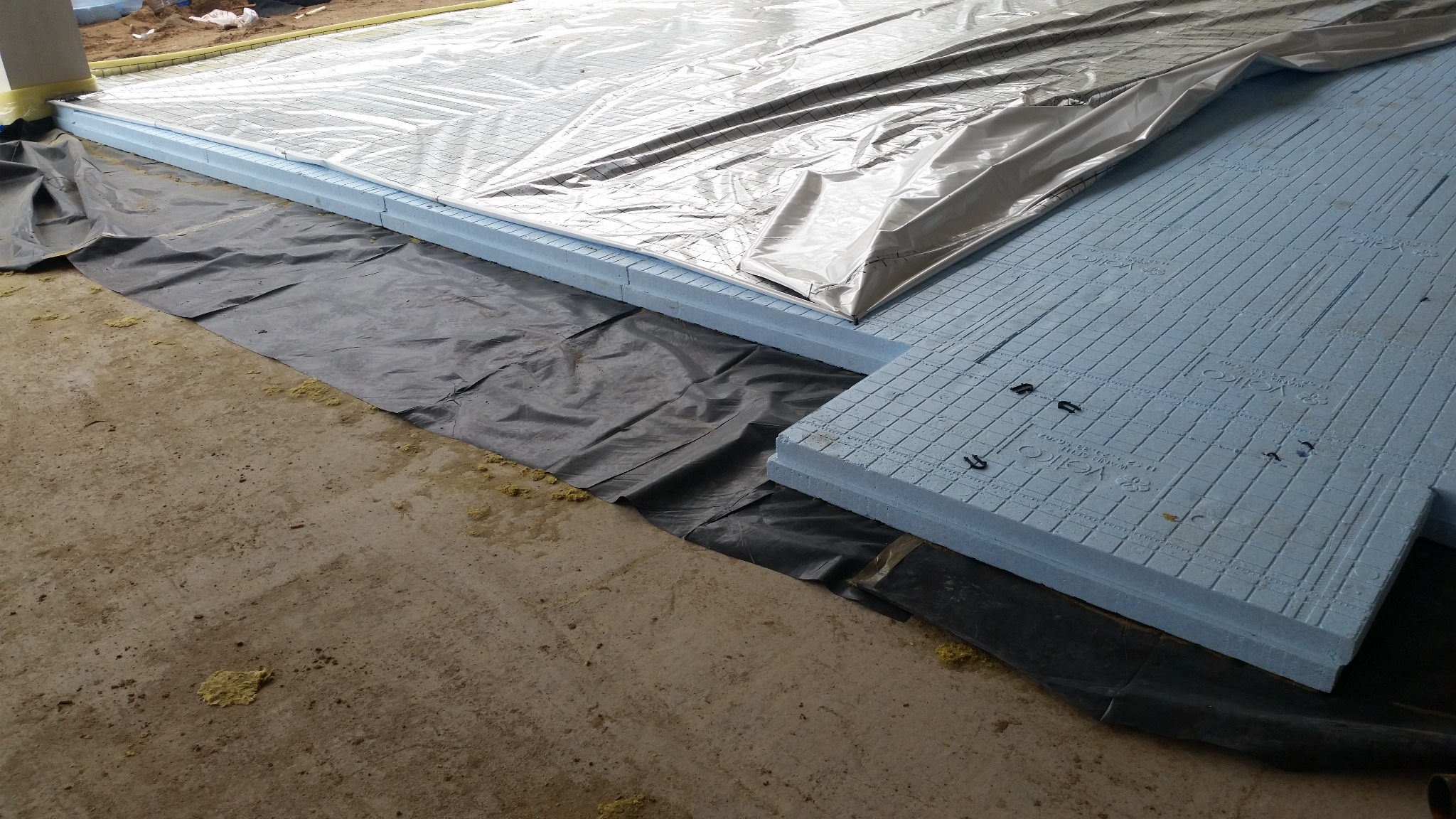 Izolacja podłogowa z płyt styropianowych na czarnej folii, przykryta folią aluminiową. Widoczne spinki mocujące. Etap prac budowlanych.