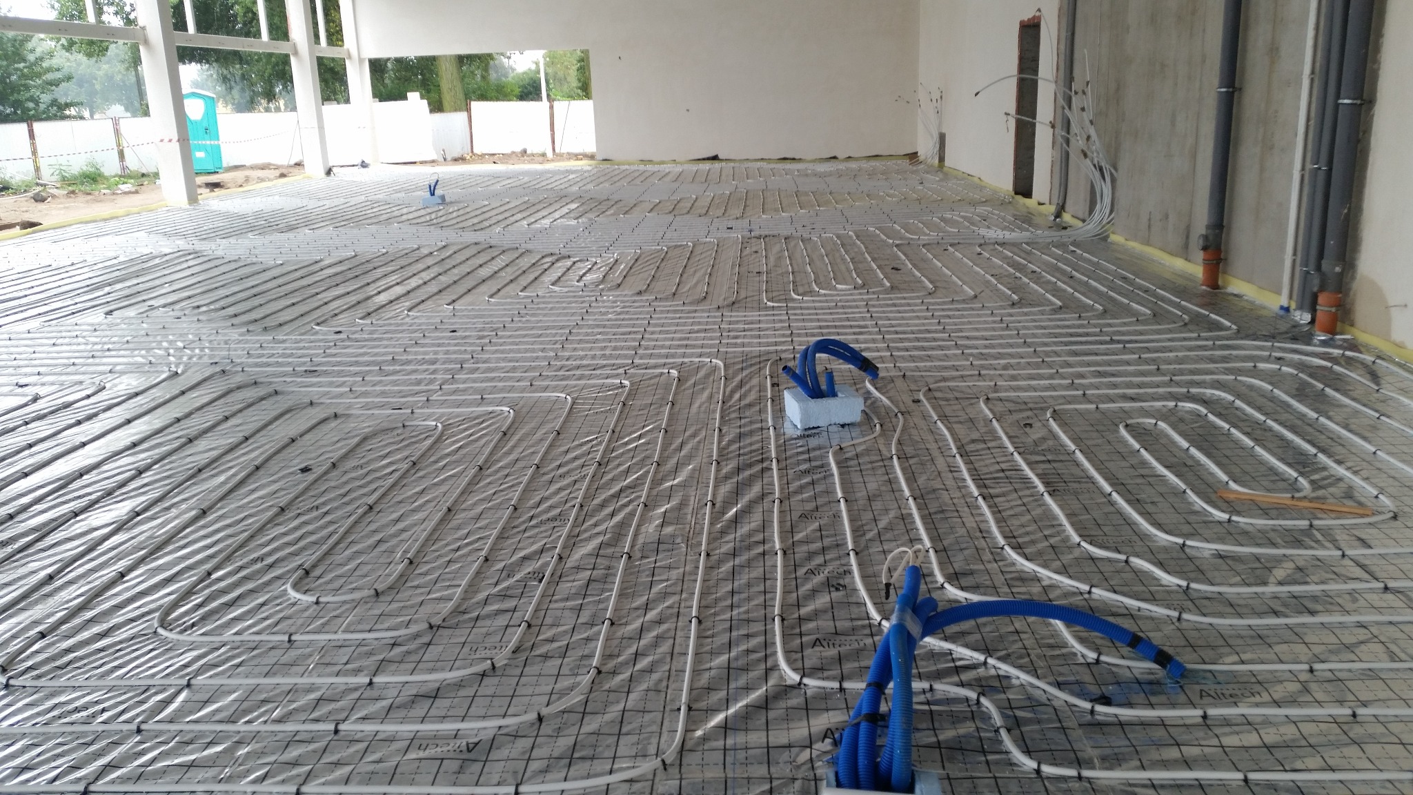 Rozległa instalacja ogrzewania podłogowego z białych rur na folii z siatką, widoczne niebieskie węże przyłączeniowe i fragmenty ścian w surowym stanie.