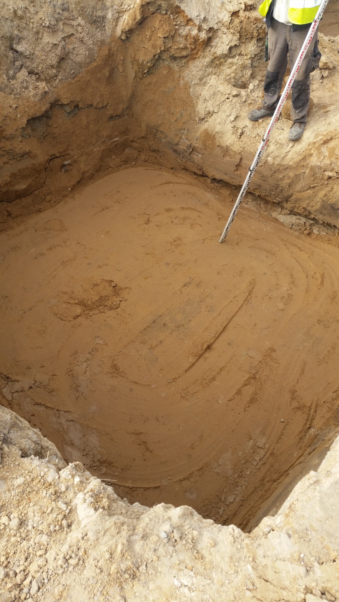 Wykop pod fundament z widocznymi śladami pracy glebogryzarki, częściowo widoczna osoba z łatą pomiarową. Grunt w wykopie jest wyrównany i przygotowany do dalszych prac.