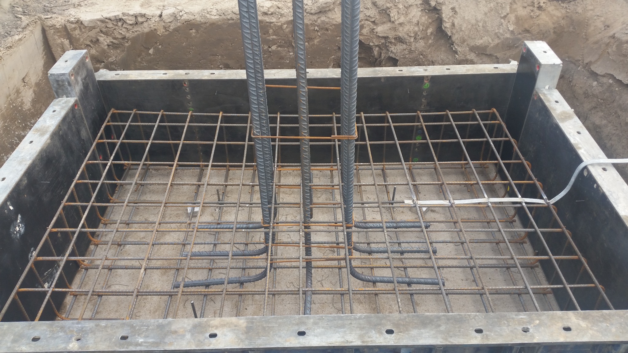 Zbrojenie fundamentu w szalunku z widocznymi pionowymi prętami i siatką zbrojeniową. Detal budowy, betonowa podstawa z elementami stalowymi.