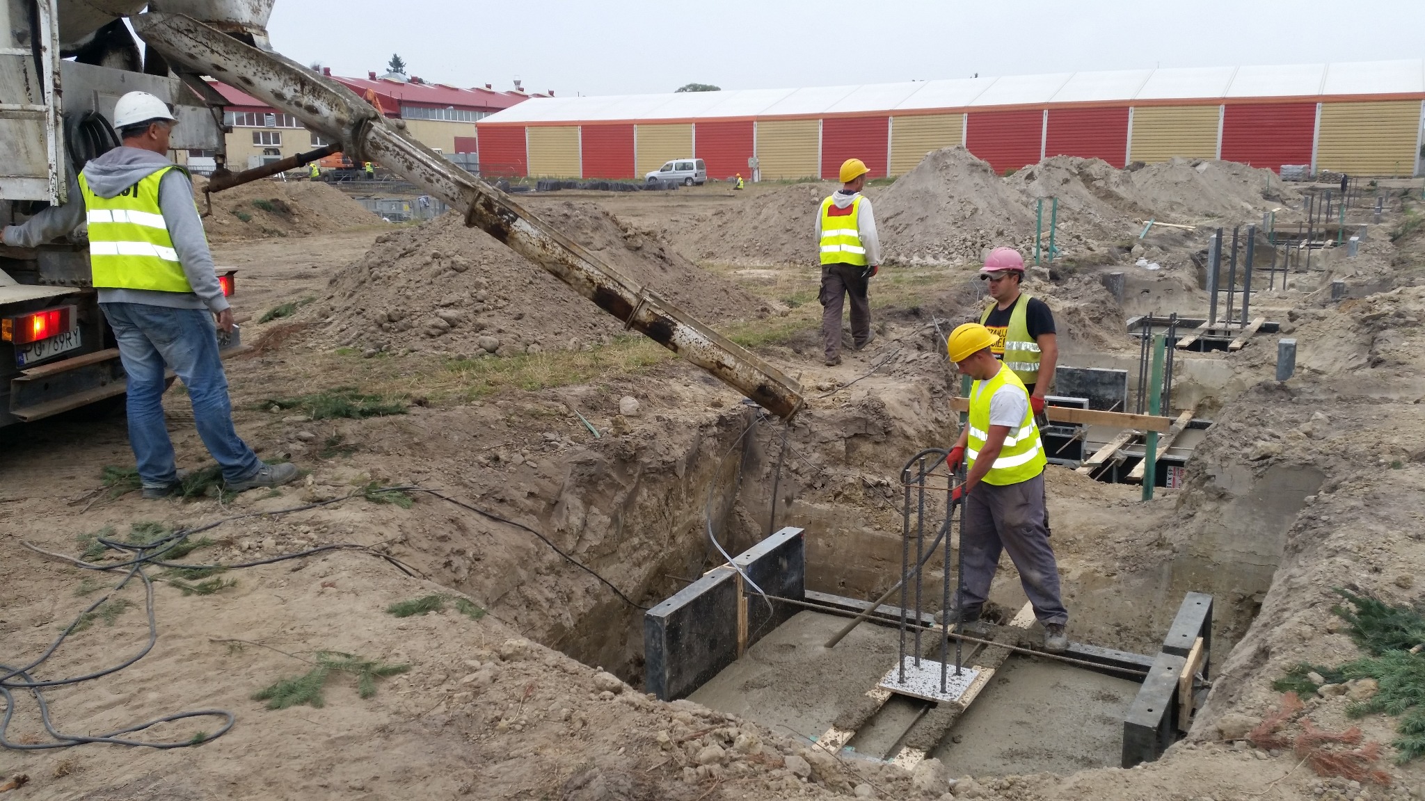 Wylewanie fundamentów pod budynek z użyciem betonomieszarki. Pracownicy w żółtych kamizelkach nadzorują proces. Widoczne zbrojenia i szalunki.