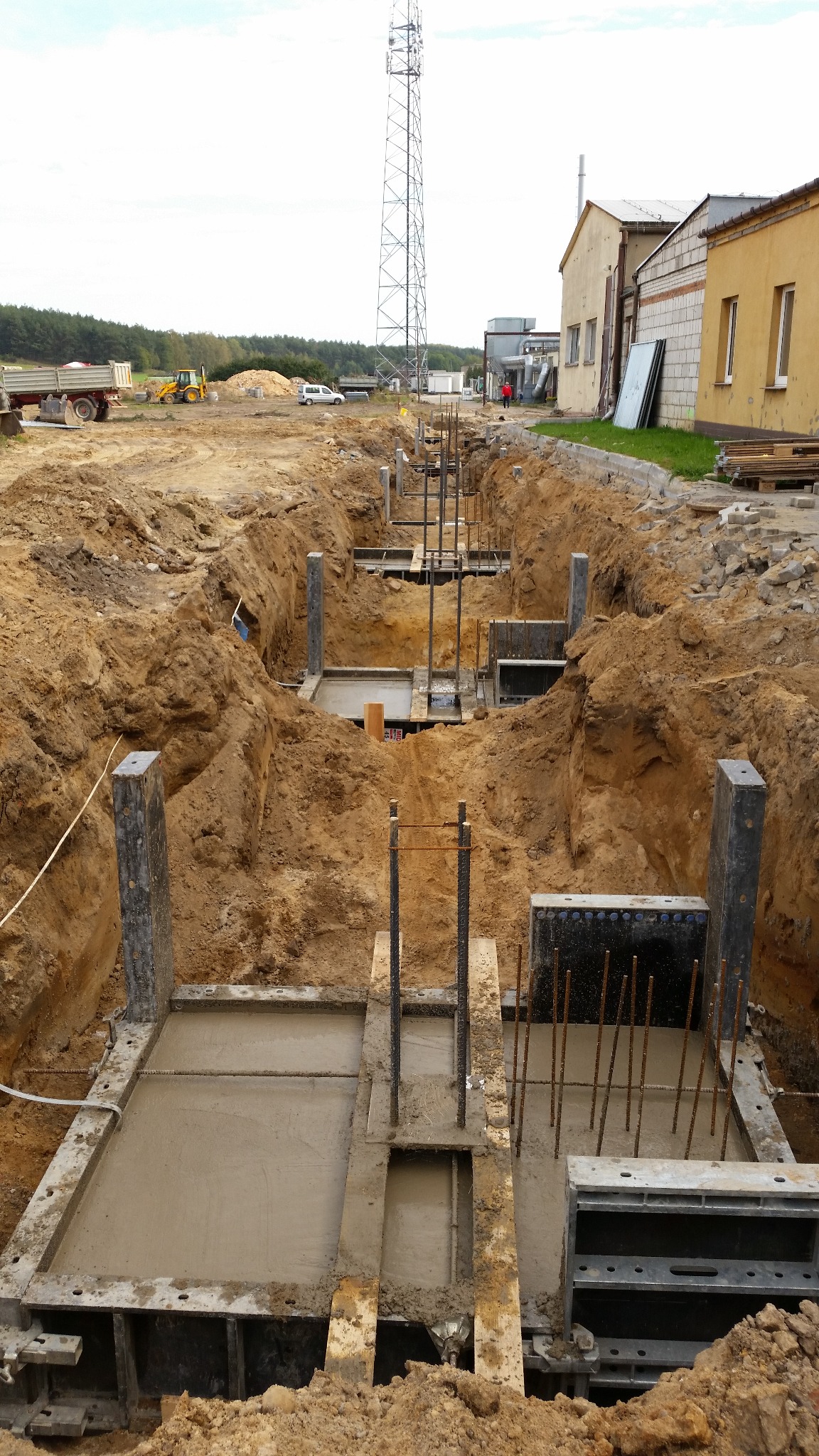Wykop z fundamentami pod budowę hali z widocznym zbrojeniem, częściowo wypełniony betonem. W tle budynek przemysłowy i wieża telekomunikacyjna.