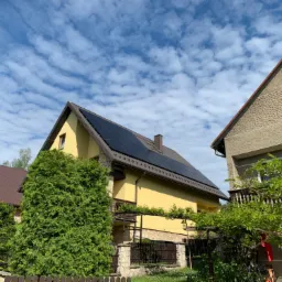 Żółty dom jednorodzinny z ciemnymi panelami na dachu, widok z poziomu gruntu, błękitne niebo z chmurami w tle.