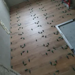 Układanie podłogi z płytek imitujących drewno z użyciem klipsów poziomujących, widoczna ściana z pianką montażową i fragment stelaża podtynkowego.