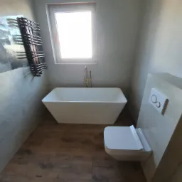 Nowoczesna łazienka z wolnostojącą białą wanną, wiszącą toaletą, grzejnikiem drabinkowym na ścianie z motywem górskim i drewnopodobnymi panelami podłogowymi, oświetlona światłem dziennym z okna.