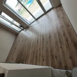 Widok z góry na nowo ułożone panele podłogowe o wzorze drewna, z dużymi oknami dachowymi w tle, widać fragment stopy w szarej skarpecie na surowym podłożu.