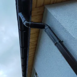 Brązowa rynna spustowa zamontowana na jasnej elewacji domu, widok z dołu na tle pochmurnego nieba.