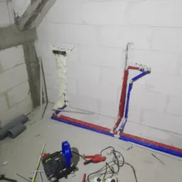 Wewnętrzne ściany z betonu komórkowego z bruzdami wypełnionymi pianką montażową i instalacją wodną z rur PEX w kolorze czerwonym i niebieskim, na podłodze leżą narzędzia: młotowiertarka, zaciskarka...
