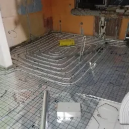 Układanie białych rur ogrzewania podłogowego na folii z nadrukiem KELLER w surowym wnętrzu, widoczne narzędzia i materiały instalacyjne.