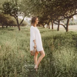 Kobieta w białej sukience spaceruje po łące z drzewami, trzymając słomkowy kapelusz, profil, ciepłe światło.