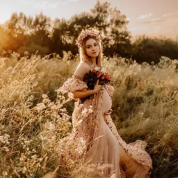 Kobieta w ciąży w zwiewnej sukni z kwiatowym wzorem i wiankiem na głowie, pozuje w wysokiej trawie o zachodzie słońca, trzyma bukiet kwiatów.