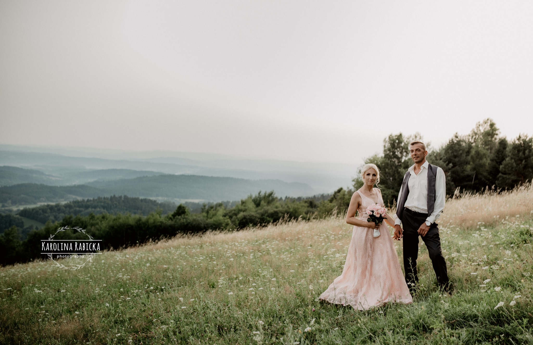 Para w eleganckich strojach, ona w pudrowej sukni, on w kamizelce, trzymająca bukiet róż, pozuje na łące z widokiem na zalesione wzgórza w oddali.