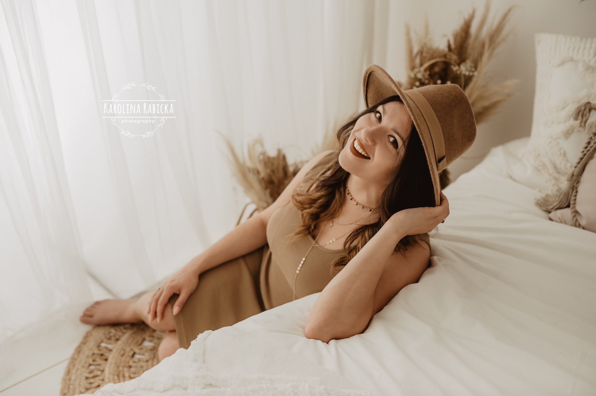 Uśmiechnięta kobieta w brązowym kapeluszu i sukience pozuje na łóżku w jasnym, naturalnym wnętrzu z dekoracjami z suszonych traw.