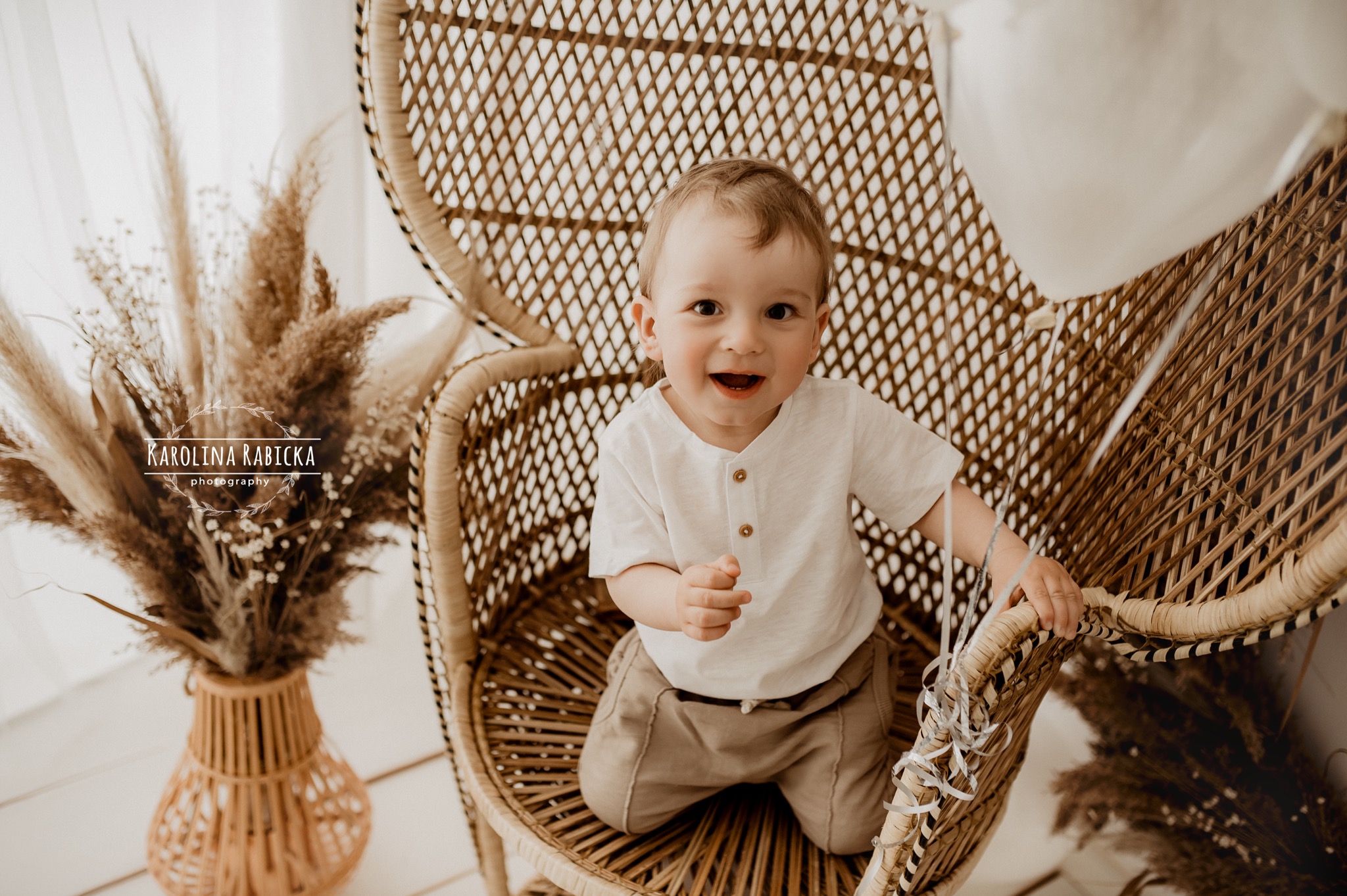 Uśmiechnięty chłopiec w beżowych ubraniach siedzi w wiklinowym fotelu z białym balonem, w tle wazon z suszonymi trawami i logo fotografa.