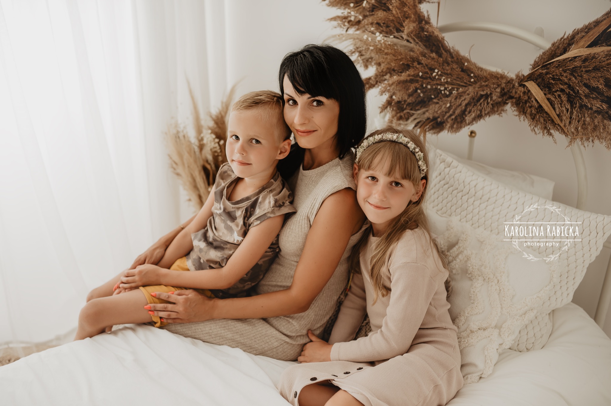 Portret matki z dwójką dzieci na łóżku z dekoracjami z suszonych traw, w jasnej tonacji kolorystycznej, wykonany w studio.