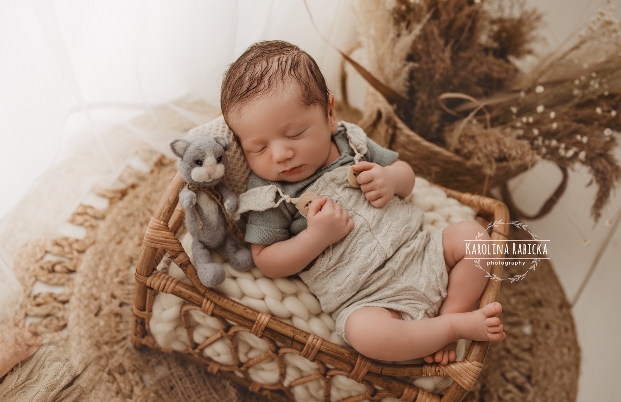 Śpiący noworodek w plecionym koszu, ubrany w dzianinowy kombinezon, obok szary pluszowy kot, w tle beżowe trawy pampasowe.