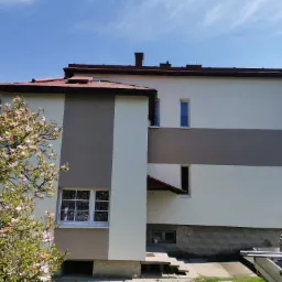 Dwukondygnacyjny dom z nową elewacją w odcieniach bieli i szarości, z kwitnącą magnolią na pierwszym planie i fragmentem rusztowania w tle.