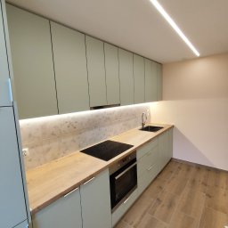 KR New Home - Nowoczesna kuchnia z jasnymi, pastelowymi szafkami, drewnianym blatem, indukcyjną płytą grzewczą i podświetleniem LED podszafkowym oraz sufitowym, wykończona płytkami imitującymi marmur.