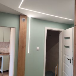 KR New Home - Korytarz z ukrytymi liniowymi lampami LED w suficie, drewnianą kolumną wentylacyjną i wejściem do łazienki z widoczną białą, częściowo otwartą, frezowaną  drzwi.