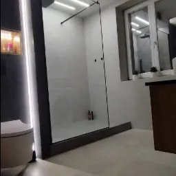 Nowoczesna łazienka z kabiną prysznicową typu walk-in, oświetleniem LED, szarą armaturą i minimalistycznym wystrojem, widok na fragment toalety i blatu z roślinami.