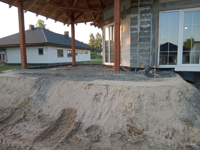 Przygotowanie podłoża pod taras z obrzeżami z kostki brukowej przy nowym budynku z widocznym poziomem i śladami opon na piasku.