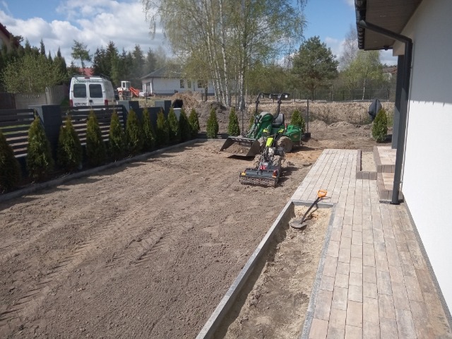 Przygotowanie terenu pod kostkę brukową: wyrównana ziemia, mini-ładowarka z łyżką i glebogryzarką, fragment ułożonej ścieżki z kostki przy budynku, widoczna łopata.