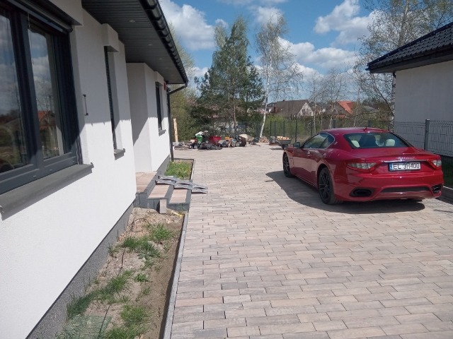 Ułożona kostka brukowa na podjeździe z czerwonym Maserati w tle, widoczny fragment elewacji domu i prace brukarskie w oddali.